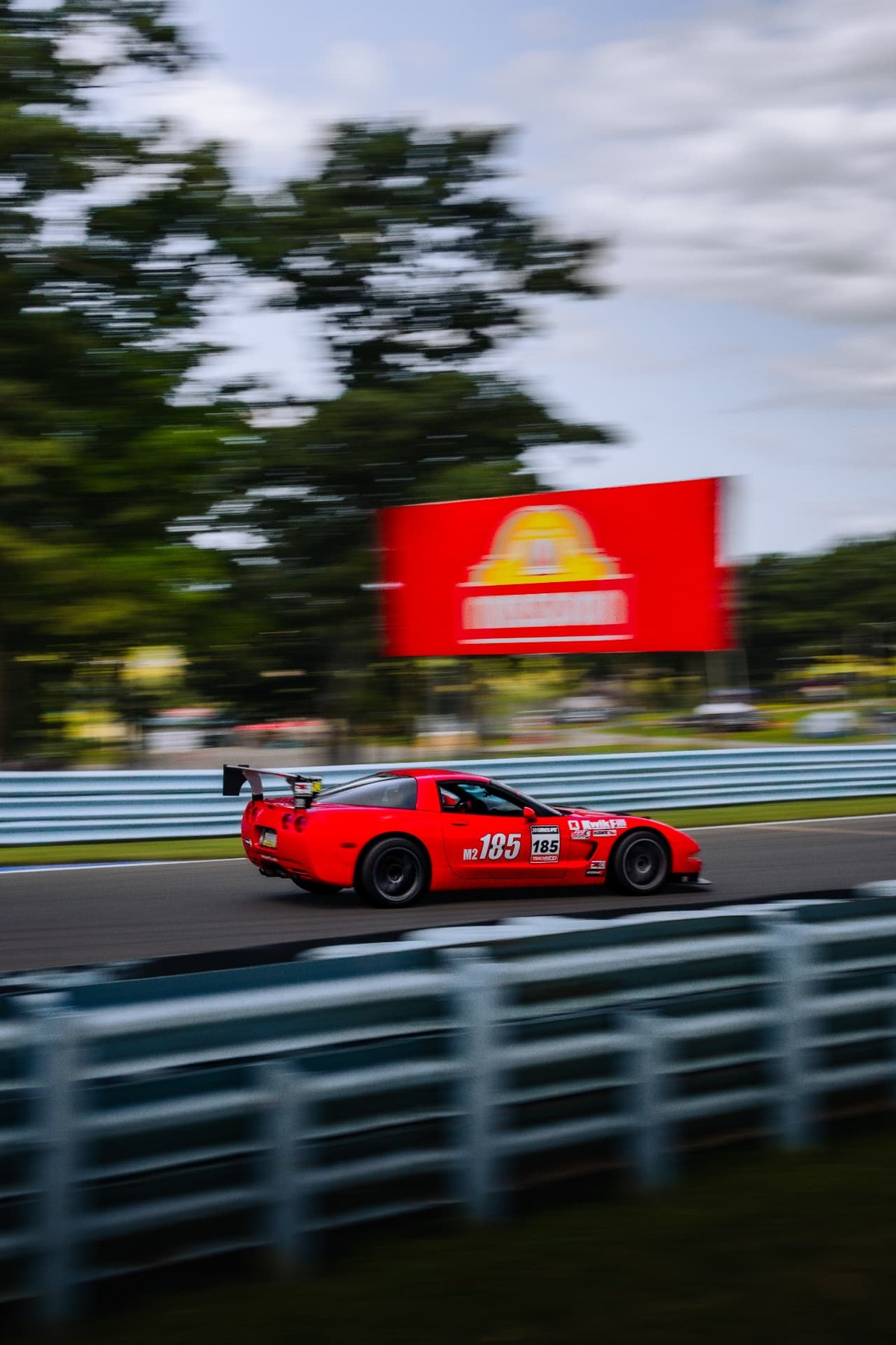 A red Corvette race car with a large rear wing, captured panning past blue guardrails at a road course